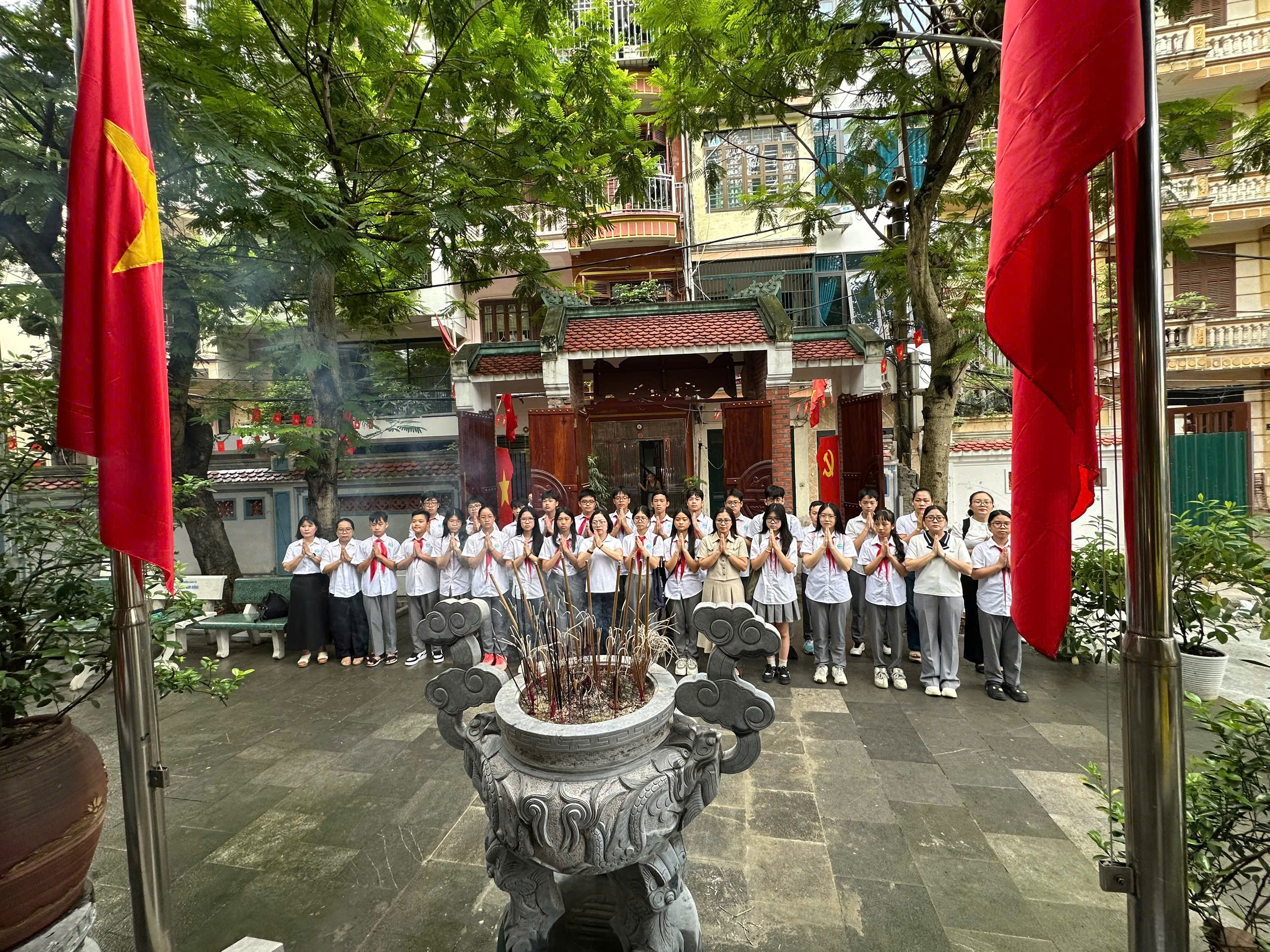 A group of people dressed in uniforms stands around a decorative fountain, with colorful buildings and a red flag in the background.AI-generated content may be incorrect.